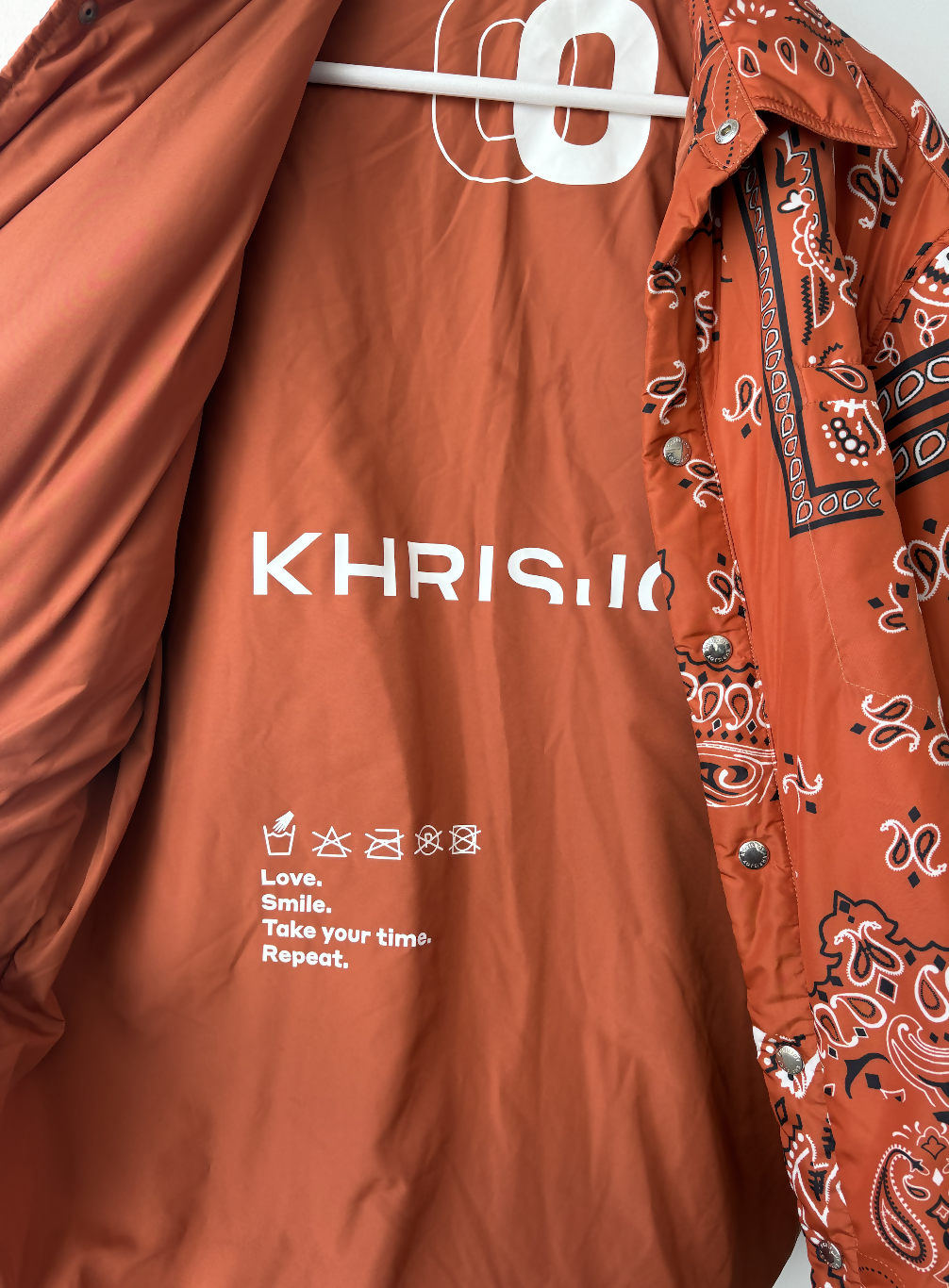 The Khrisjoy Oranga Bandana Print Shirt Jacket by EXCHANGE STORE features orange paisley-print sleeves and an open front showing the bold white KHRISJOY logo with care instructions: Love. Smile. Take your time. Repeat.
