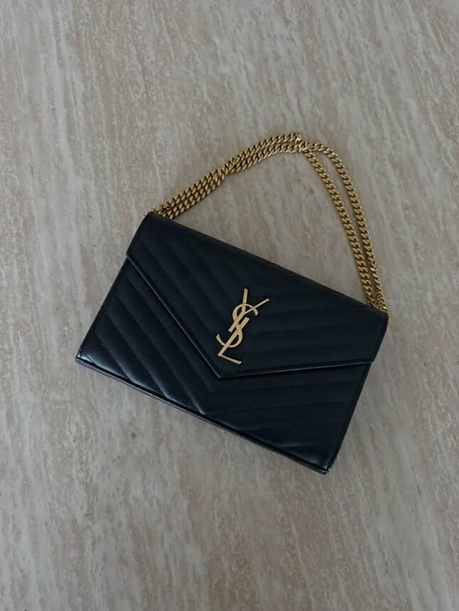 A Saint Laurent Classic Cassandre chain wallet in grain de poudre leather from EXCHANGE STORE, featuring a gold chain strap and signature YSL logo, displayed on a light-colored textured surface.
