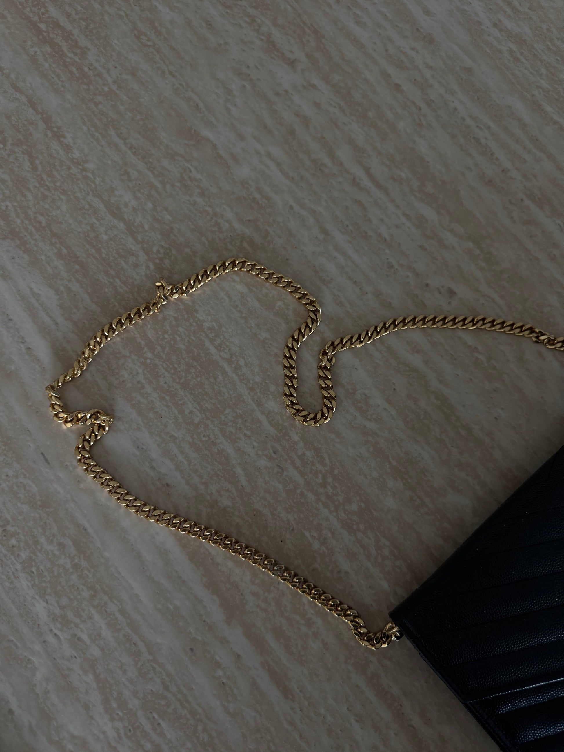 A gold chain is attached to the corner of the EXCHANGE STORE Saint Laurent Classic Cassandre chain wallet in grain de poudre black leather with chevron quilting, resting on a beige textured surface.