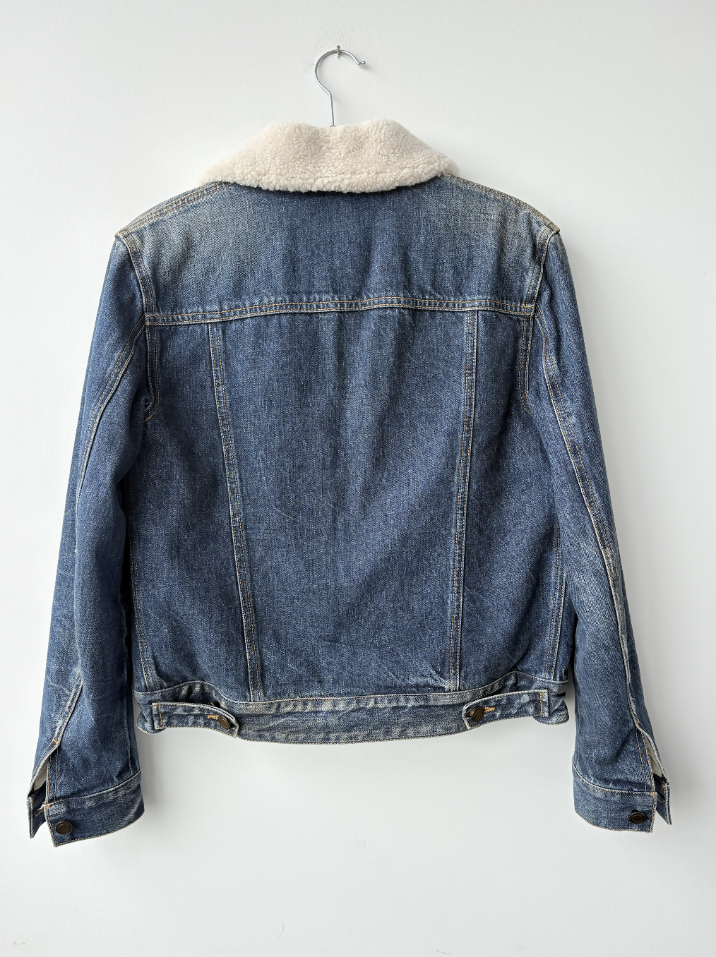 The Yves Saint Laurent Paris Denim Shearing Jacket by EXCHANGE STORE, featuring blue denim and a light sherpa collar, hangs on a metal hanger against a white wall, shown from the back.