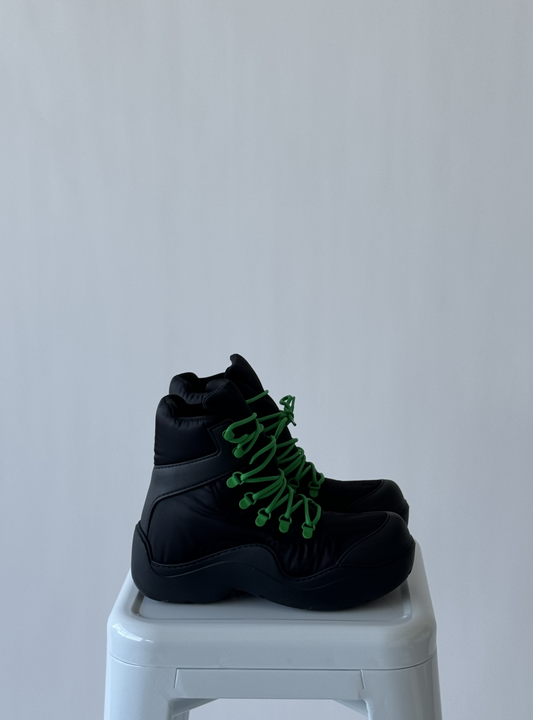 A pair of black Bottega Veneta Puddle Lace Up Boots by EXCHANGE with bright green laces, size 38, is displayed side by side on a white stool against a plain light gray background.
