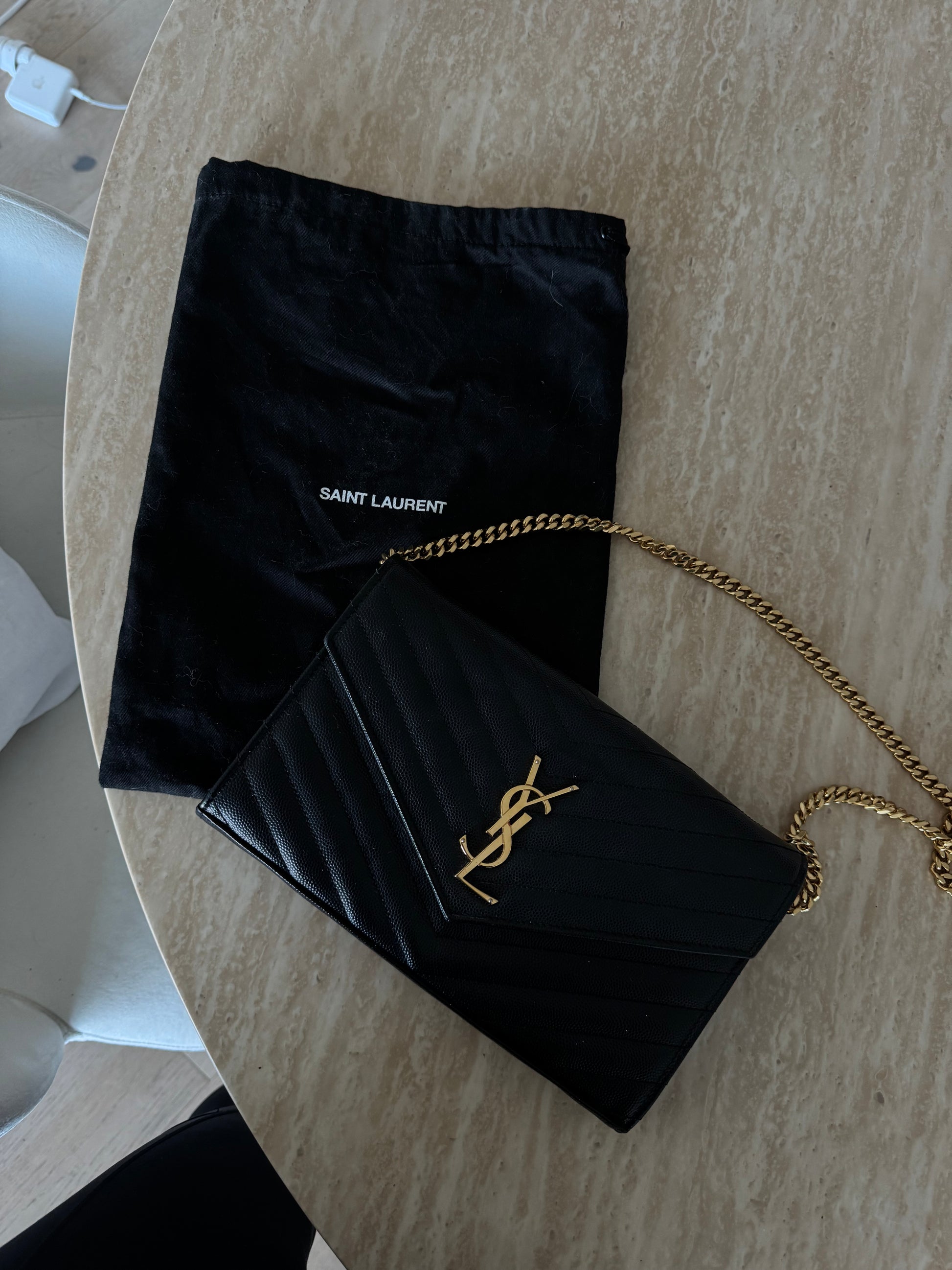 The Saint Laurent Classic Cassandre chain wallet in grain de poudre leather from EXCHANGE STORE, with gold YSL logo and chain, is displayed on a marble surface beside its matching dust bag.