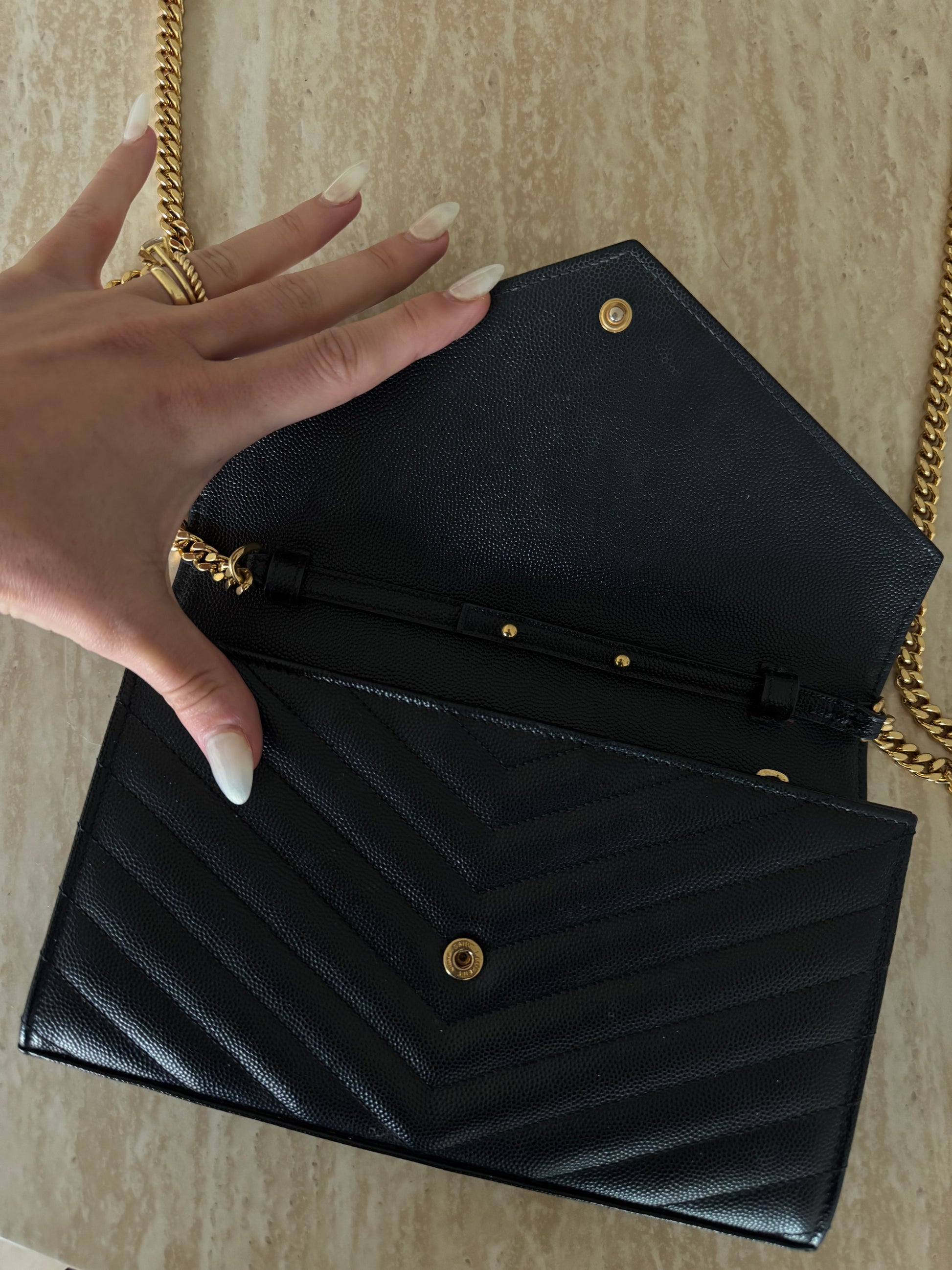 A hand with manicured nails and gold rings opens the EXCHANGE STORE Saint Laurent Classic Cassandre chain wallet in grain de poudre leather on a light marble surface.