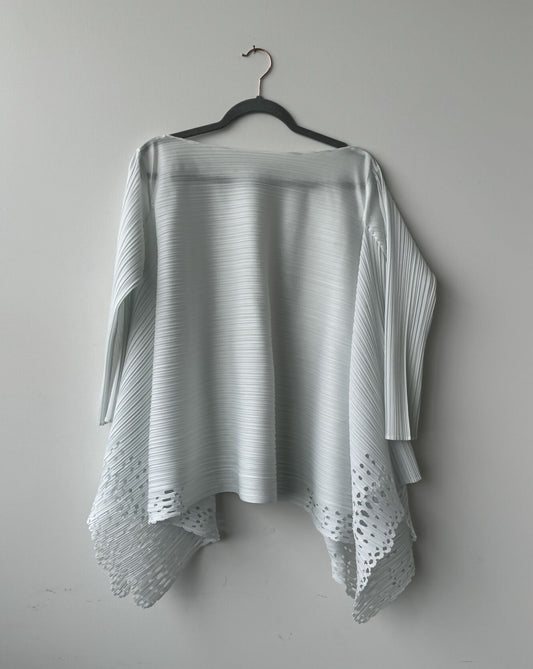 An Issey Miyake Pleats Please Top by EXCHANGE, in light gray with laser-cut floral details and an asymmetrical hem, is displayed on a black hanger against a plain light gray wall.