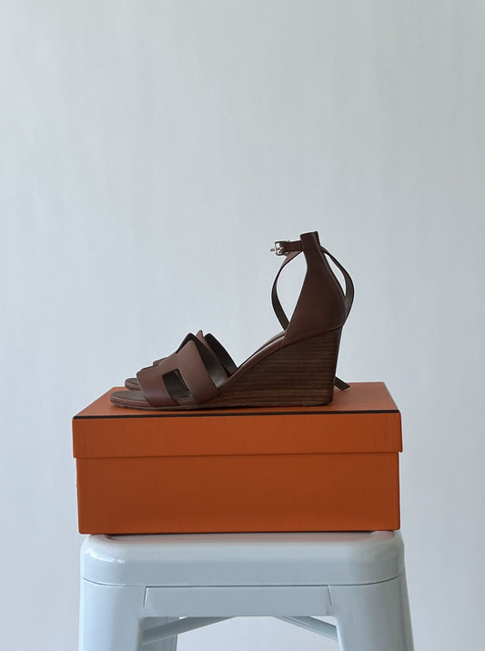 A pair of HERMES Calfskin Legend Wedge Sandals by EXCHANGE, size EU 39, in brown with ankle strap, is displayed on an orange shoebox atop a white stool against a plain light background.
