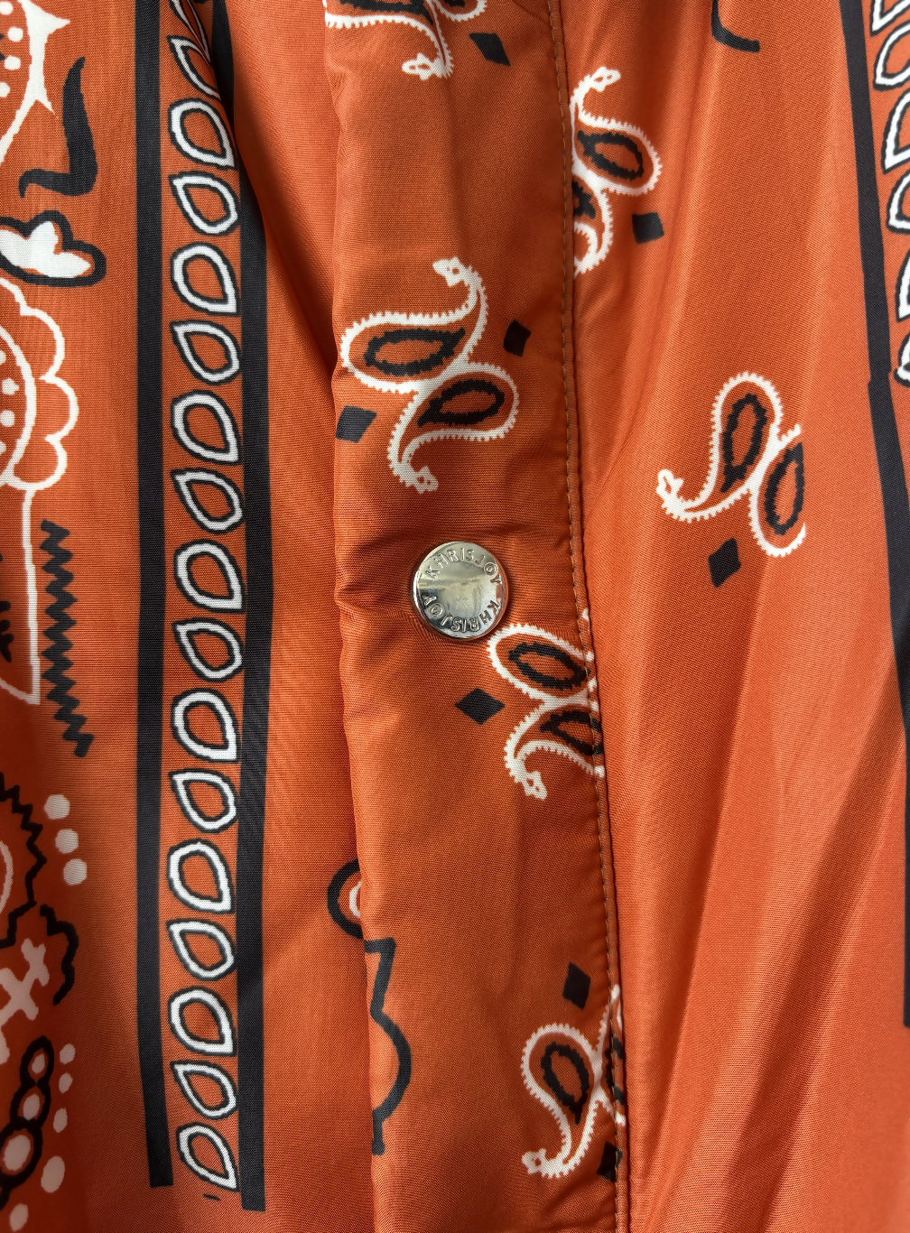 Close-up of the Khrisjoy Oranga Bandana Print Shirt Jacket by EXCHANGE STORE, showing orange fabric with black-and-white paisley and geometric patterns, plus a silver CLASSIC JEANS snap button—echoing an oversized silhouette.