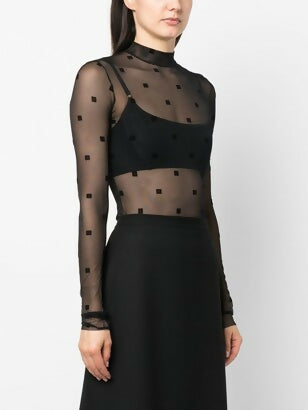 A woman wears the Givenchy Logo Print Black Mesh Bodysuit by EXCHANGE STORE, featuring a sheer black long-sleeve high neck with square patterns, layered over a black bra and paired with a black skirt.