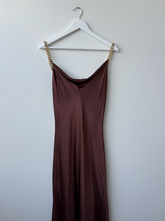 An Olivia Von Halle Bibi Silk Chain Dress from EXCHANGE STORE, in silky dark brown with gold chain straps, hangs on a white hanger against a plain white wall.