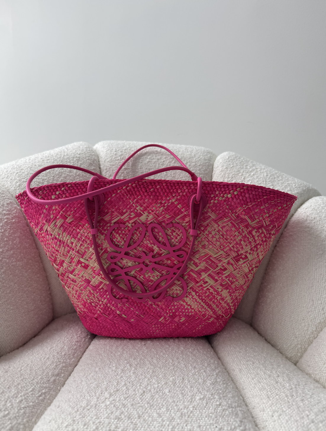 The Loewe Raffia Pink Wicker Basket Tote 2023 by EXCHANGE STORE, in excellent condition with leather handles and detailing, rests on a textured white armchair against a plain light gray wall.