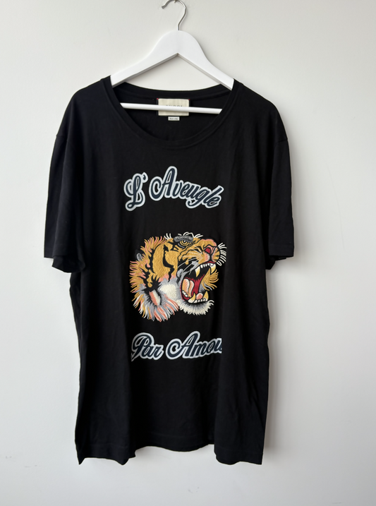 A Gucci Black L'Aveugle Par Amour T-Shirt from EXCHANGE STORE hangs on a white hanger, featuring an embroidered tiger head and “L’Aveugle Par Amour” in elegant white script.