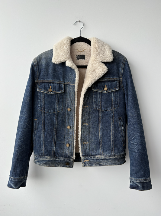 Yves Saint Laurent Paris Denim Shearing Jacket by EXCHANGE STORE, featuring blue denim with white sherpa collar and lining, buttoned front, flap breast pockets, and side pockets, displayed on a black hanger against a white wall.