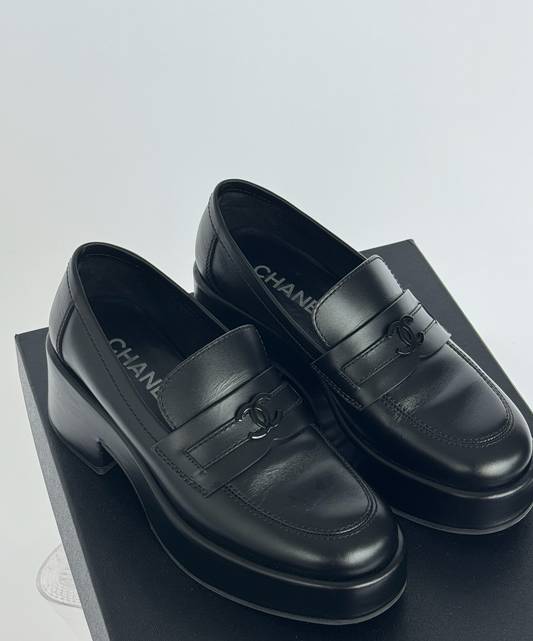 A pair of EXCHANGE STORE Chanel Moccasin Loafers - FULL SET in black calfskin with the iconic CC logo on the strap, shown atop a black shoebox against a white background, exudes understated luxury.