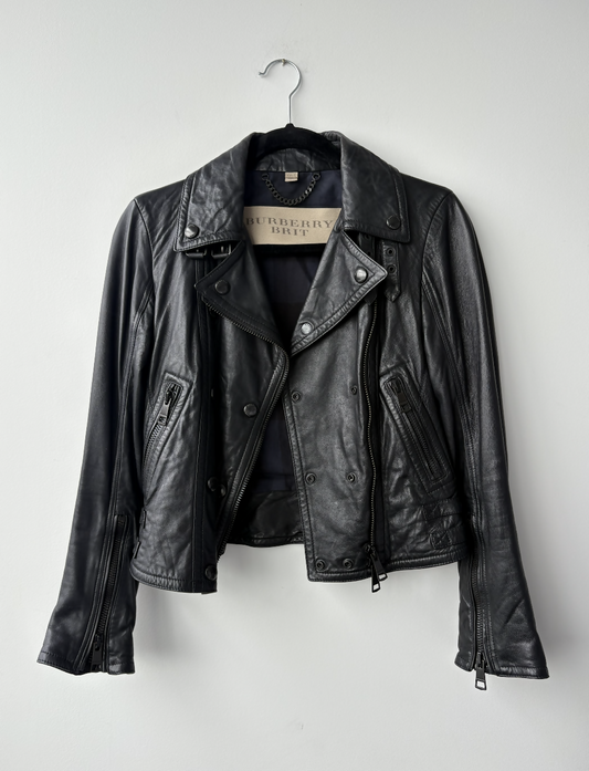 The Burberry Brit Black Leather Biker Jacket by EXCHANGE STORE hangs on a metal hanger against a white wall, showcasing zippered pockets, lapels, and the brand label inside the collar.
