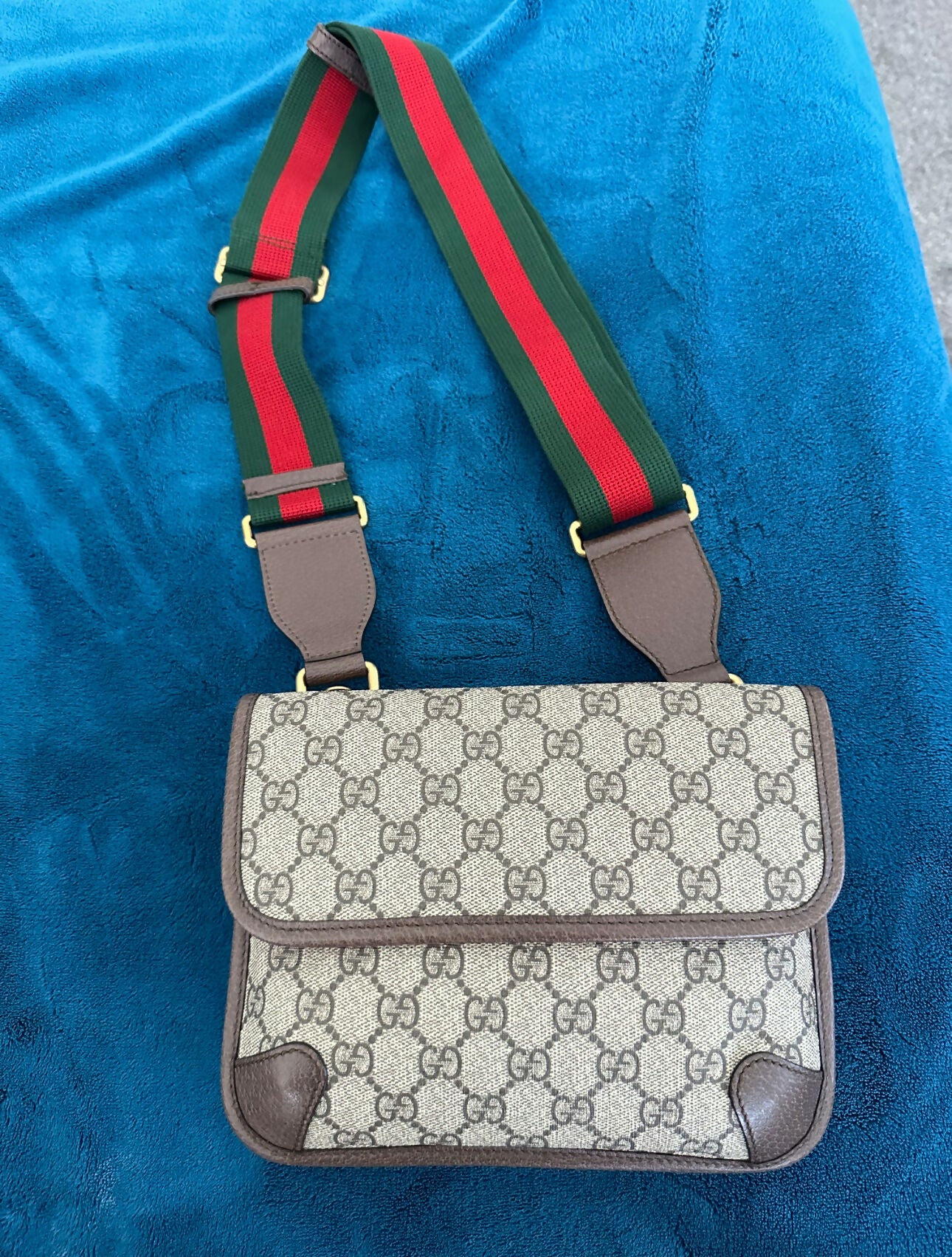 The EXCHANGE STORE Gucci Neo Vintage messenger bag with GG monogram, brown leather trim, and Web stripe strap is displayed on a bright blue fabric background.