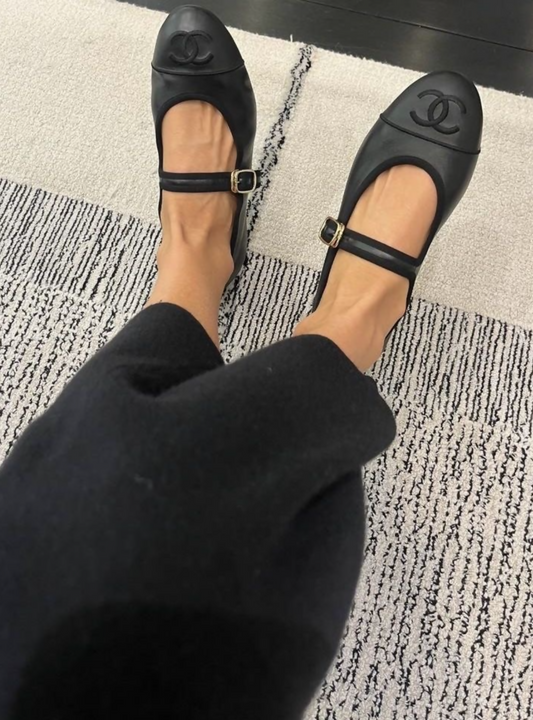 A person wearing EXCHANGE STORE Chanel Mary Jane flats in black lambskin (FULL SET) is seated on a light textured rug, with lower legs and part of dark clothing visible—a must-have for Chanel collectors.