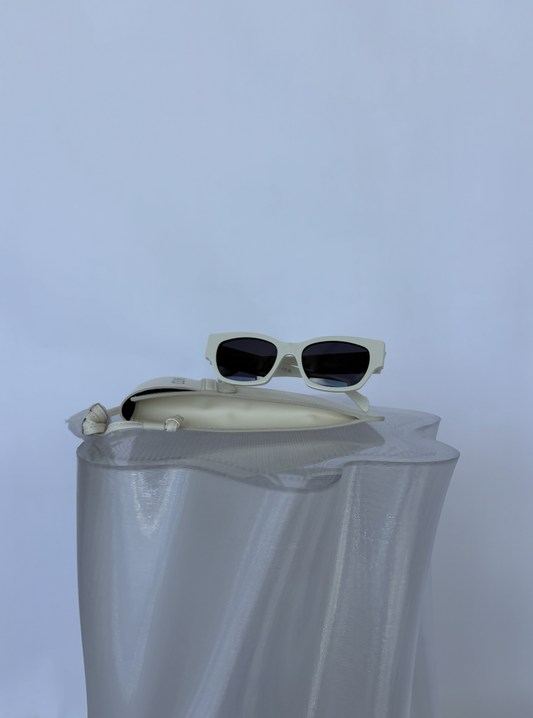 The Celine Beige Sunglasses by EXCHANGE STORE, featuring a beige frame and green lenses, rest on a white case atop a wavy reflective surface against a plain light background.