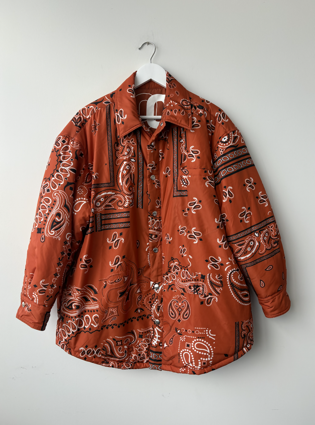The Khrisjoy Oranga Bandana Print Shirt Jacket by EXCHANGE STORE, featuring a rust bandana-inspired print and an oversized fit, hangs on a white hanger against a plain white wall.