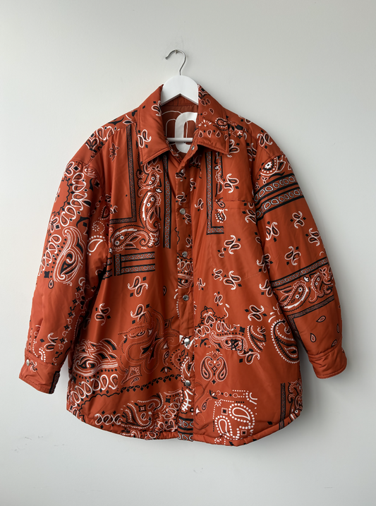 The Khrisjoy Oranga Bandana Print Shirt Jacket by EXCHANGE STORE, featuring a rust bandana-inspired print and an oversized fit, hangs on a white hanger against a plain white wall.