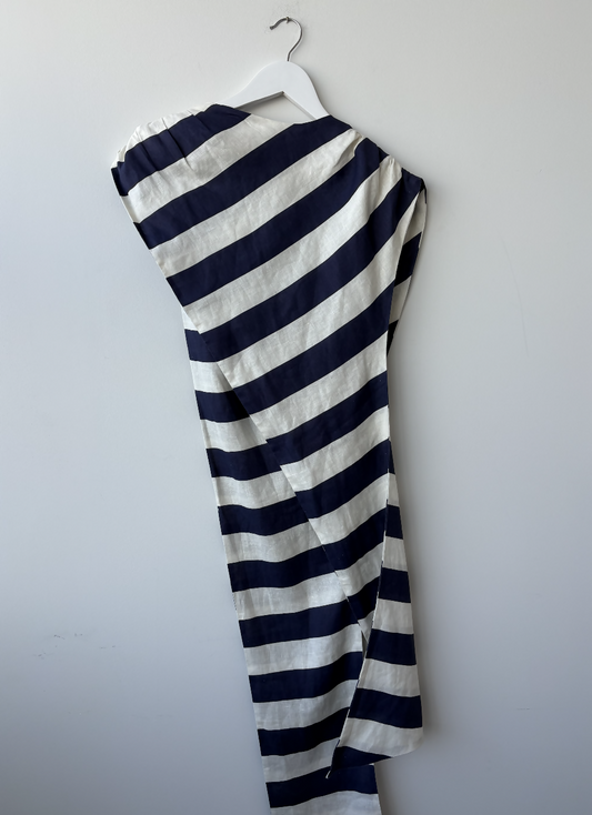 The EXCHANGE STORE Paper London Navy Stripe One Shoulder Tie Top, featuring wide navy and white stripes in an asymmetrical draped design, is displayed on a white hanger against a plain light gray wall.