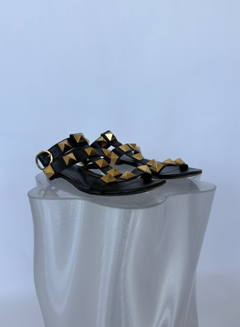 A pair of Valentino Garavani Roman Stud Sandals Black - FULL SET by EXCHANGE STORE, with bold gold pyramid studs on black leather straps, displayed on a wavy translucent pedestal against a simple light background.