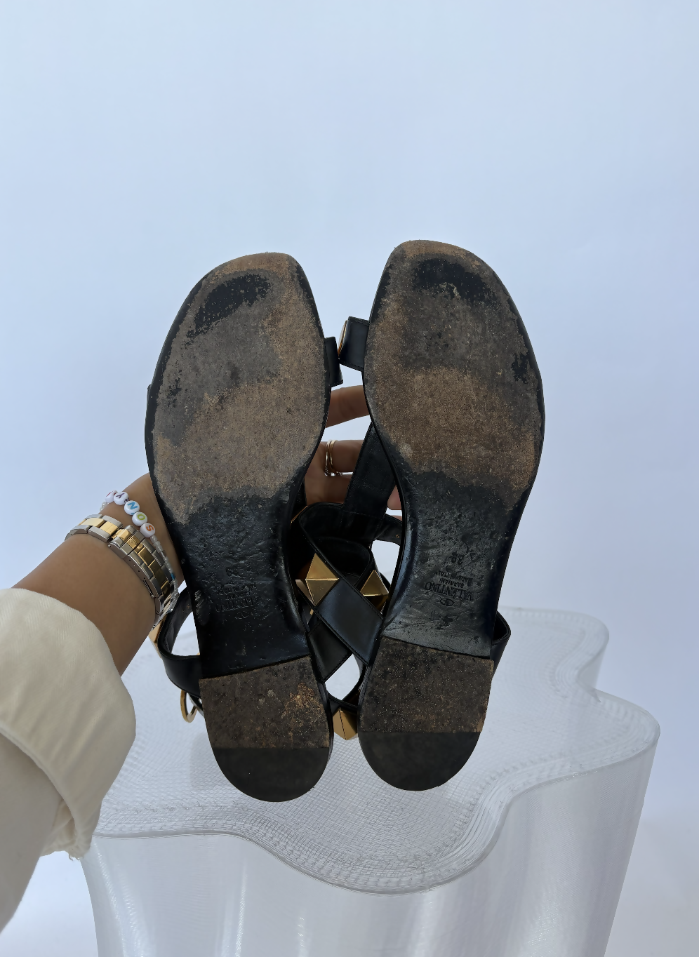 A person in a cream sleeve and bracelets holds up EXCHANGE STORE’s Valentino Garavani Roman Stud Sandals Black - FULL SET, displaying the worn, scuffed black leather soles against a plain light background.
