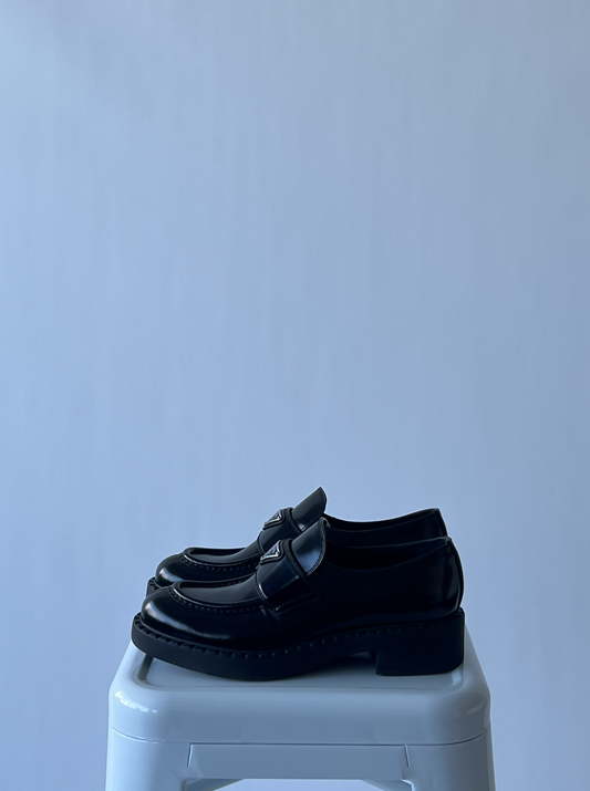 A pair of size 38.5 EXCHANGE Prada Loafers - Full Set in black with chunky soles rests on a white stool against a light blue background.