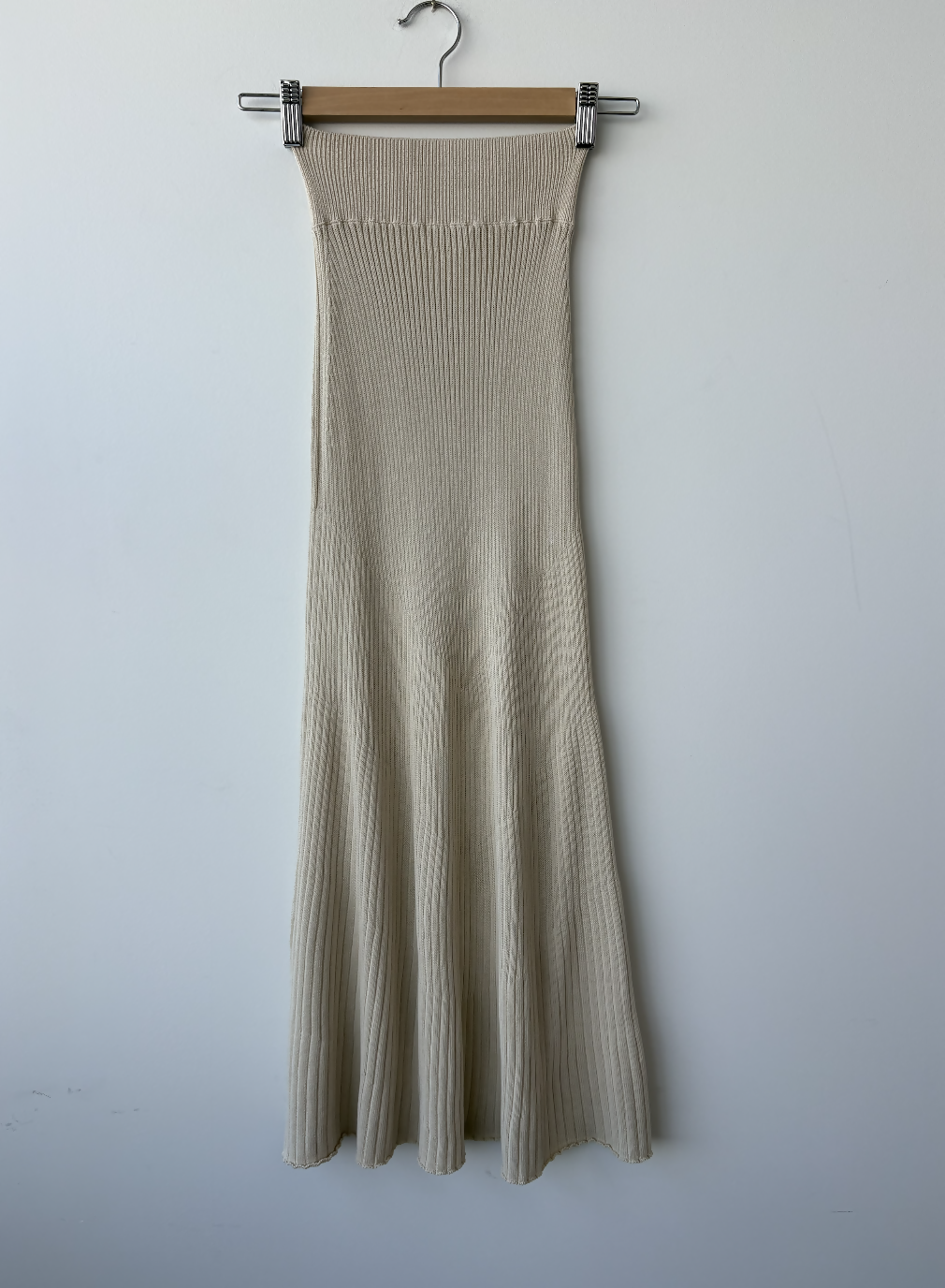 The Dadelszen Beiged Ribbed Knit Skirt by EXCHANGE STORE, featuring a high fitted waist and flared hem, hangs on a wooden hanger against a plain white wall—an ideal addition to any luxury wardrobe.