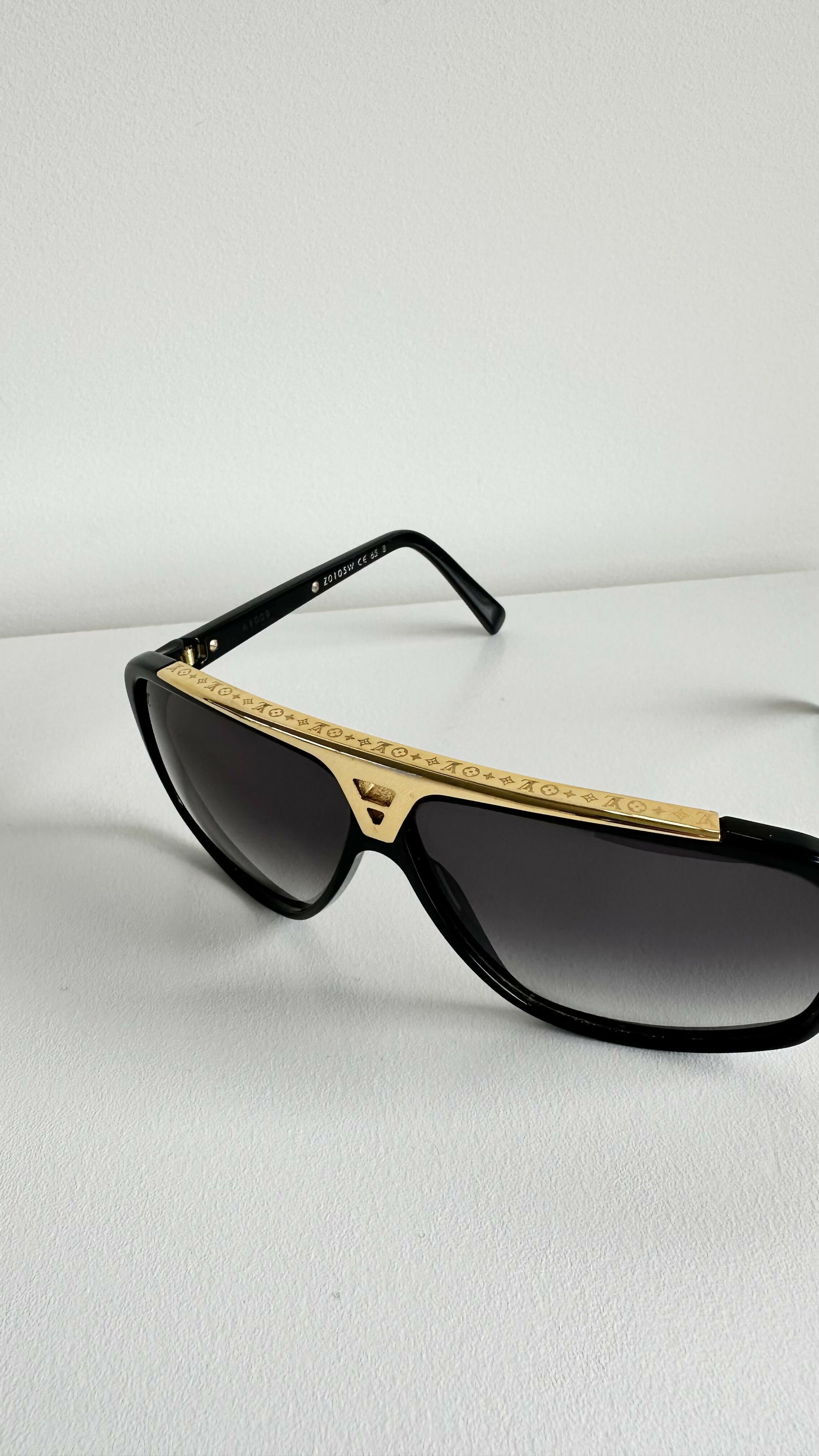 A pair of Louis Vuitton Evidence Noir W Sunglasses by EXCHANGE STORE, featuring a black frame with a gold top accent and dark lenses, displayed on a white surface against a plain white background.