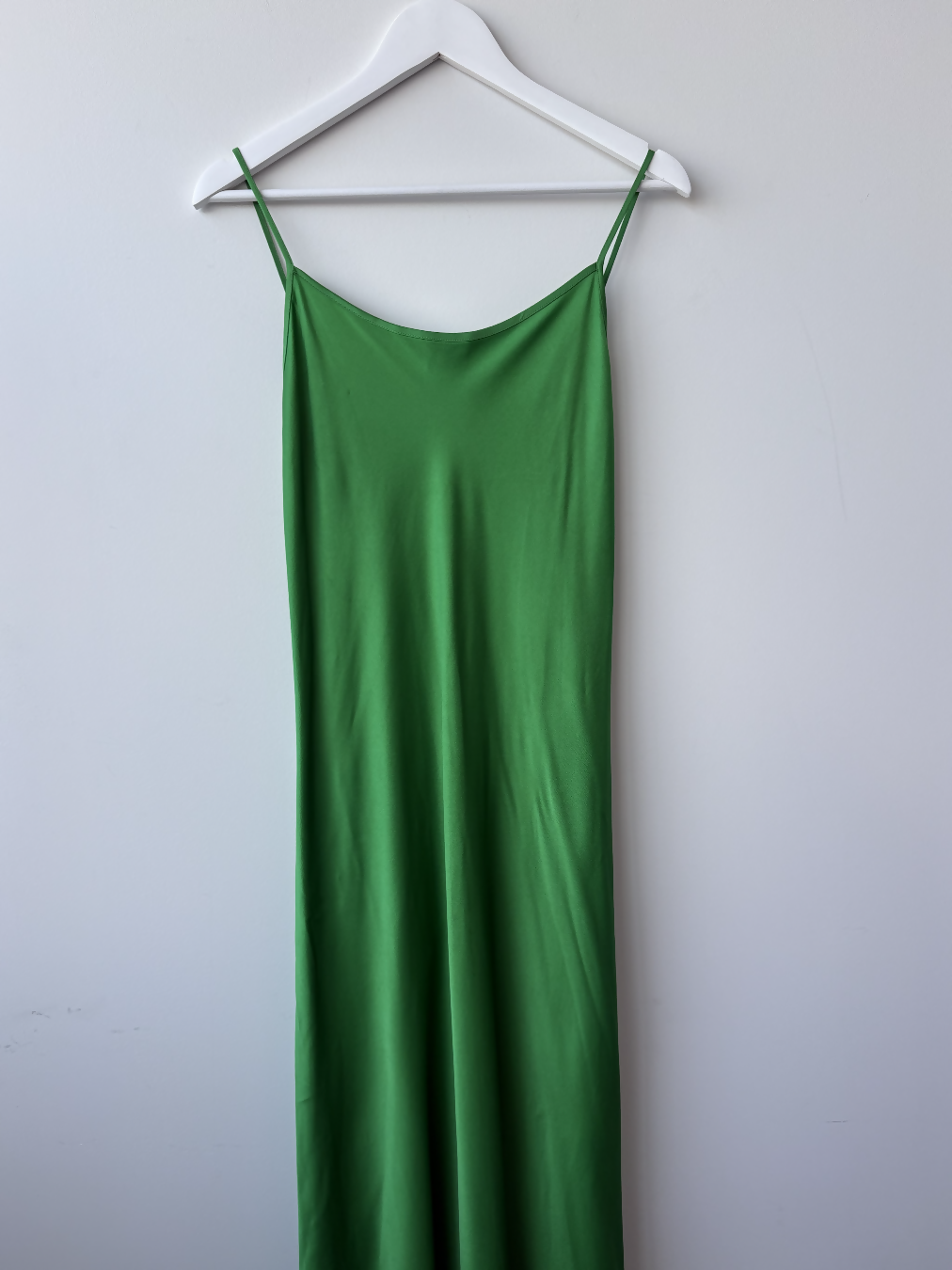 The Victoria Beckham Resort 2022 Green Cami Slip Dress by EXCHANGE STORE hangs sleeveless with thin straps on a white hanger against a light gray wall, exuding effortless elegance.