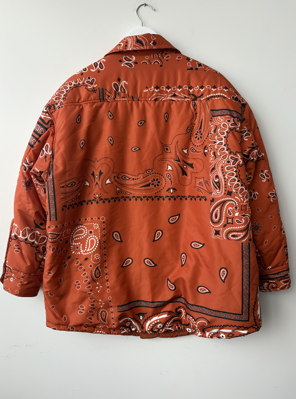 The Khrisjoy Oranga Bandana Print Shirt Jacket by EXCHANGE STORE, featuring a rust color and white/black paisley print, hangs back-facing on a wall-mounted hook.