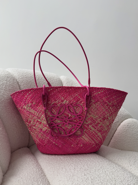 The Loewe Raffia Pink Wicker Basket Tote 2023 by EXCHANGE STORE, featuring pink and beige patterns, pink leather handles, and a pink leather logo, is displayed on a light textured armchair.