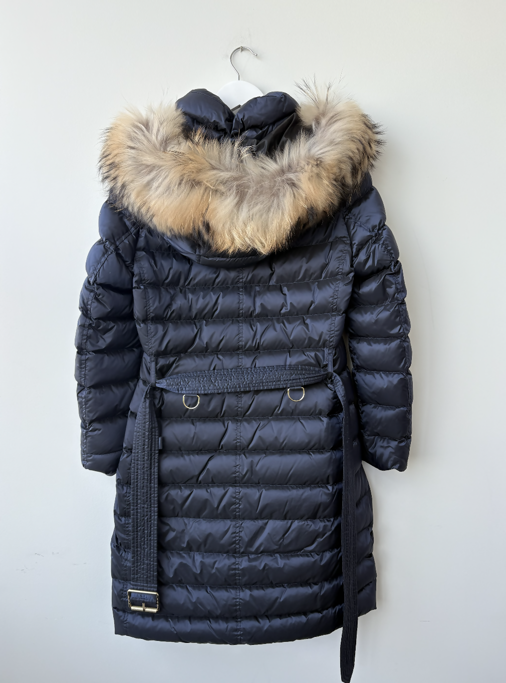 The EXCHANGE STORE Burberry Puffer with Fur Hood, featuring a dark quilted design, fur-trimmed hood, belted waist, and metallic buckle details, hangs on a white wall for the ultimate luxury winter style.