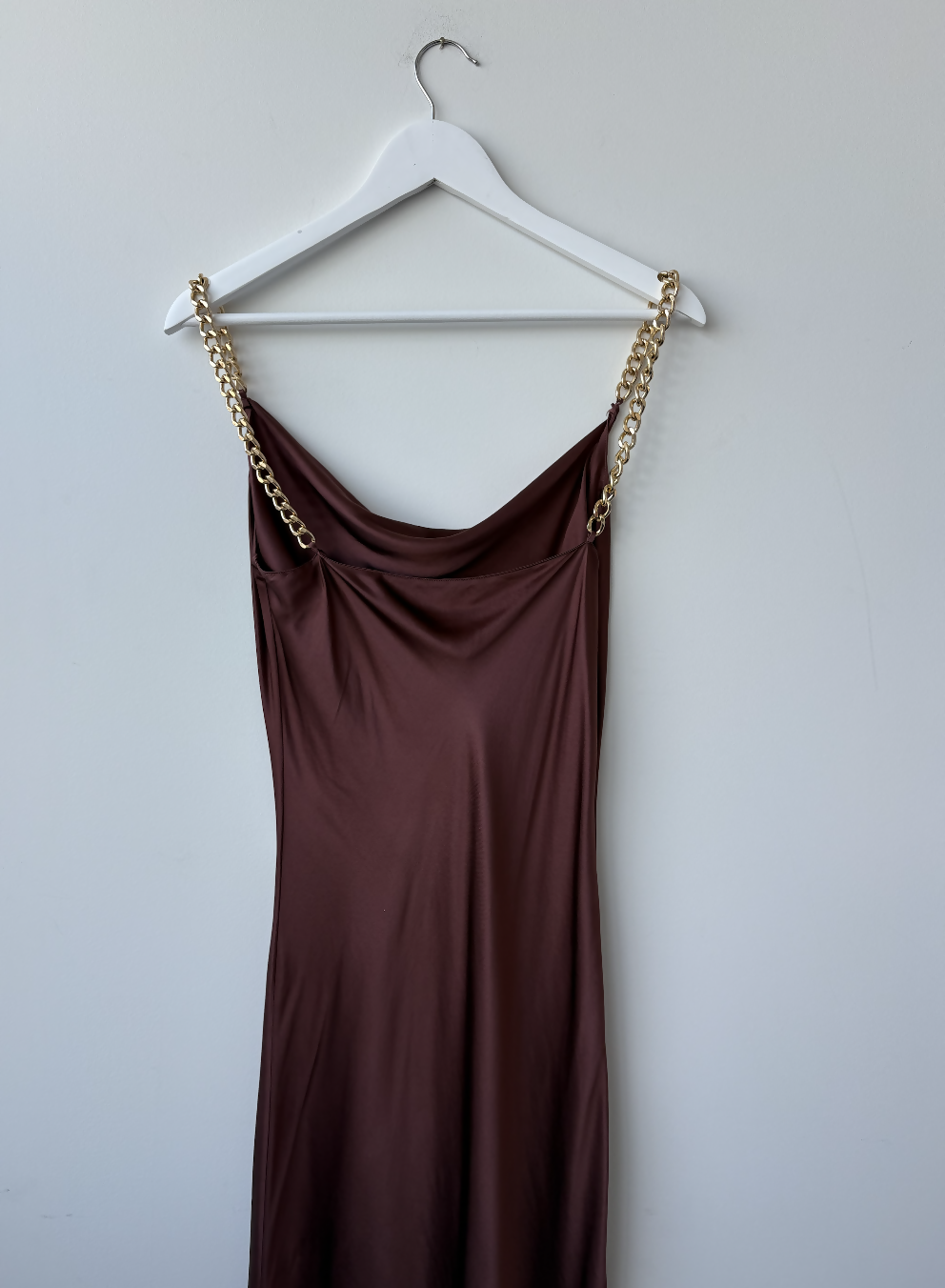The Olivia Von Halle Bibi Silk Chain Dress by EXCHANGE STORE is a sleeveless brown silk slip dress with a draped neckline and gold chain straps, displayed on a white hanger against a plain white wall.