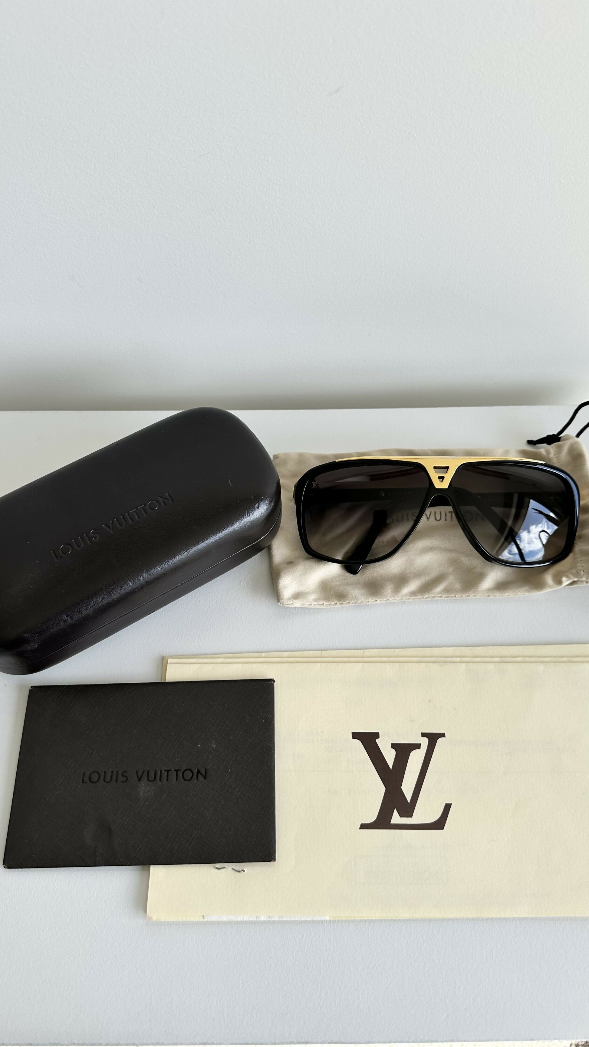 A pair of Louis Vuitton Evidence Noir W Sunglasses from EXCHANGE STORE sits on a cloth, next to a black case, black booklet, and cream card with the LV logo. All items rest on a white surface.