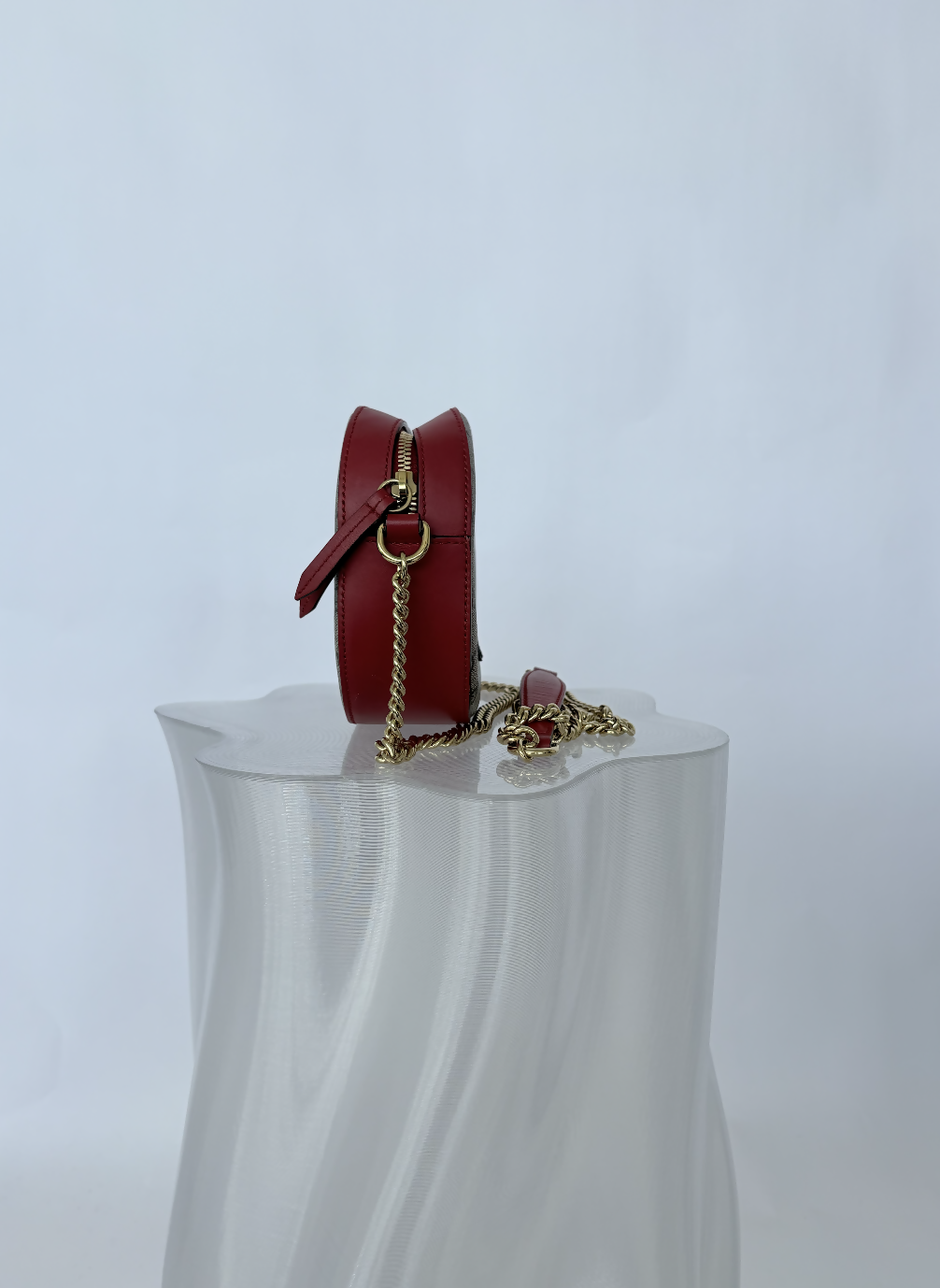 The Gucci Kingsnake Heart Mini GG Supreme Chain Crossbody Bag by EXCHANGE STORE, featuring red leather and a gold chain strap, is displayed on a white textured pedestal against a plain white background.