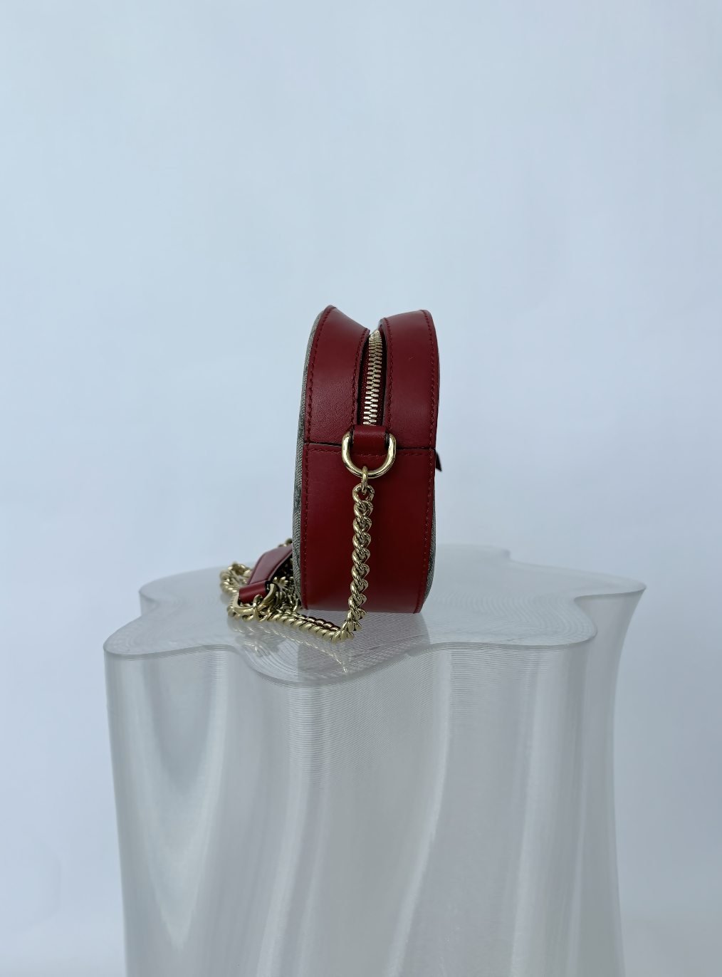 The Gucci Kingsnake Heart Mini GG Supreme Chain Crossbody Bag by EXCHANGE STORE, featuring a gold chain strap and zipper, is displayed upright on a translucent wavy pedestal against a plain light background.