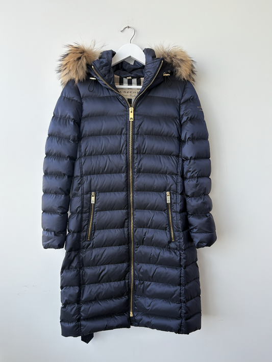 The Burberry Puffer with Fur Hood by EXCHANGE STORE is a long navy blue winter jacket featuring a fur-trimmed hood, two zippered front pockets, and a gold zipper, displayed on a white hanger against a light-colored wall.