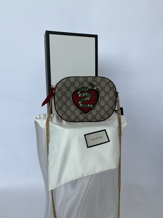 The EXCHANGE STORE Gucci Kingsnake Heart Mini GG Supreme Chain Crossbody Bag - FULL SET is shown on a GUCCI-labeled dust bag in front of a white box, featuring GG Supreme canvas with red heart and green snake details and a gold chain strap.