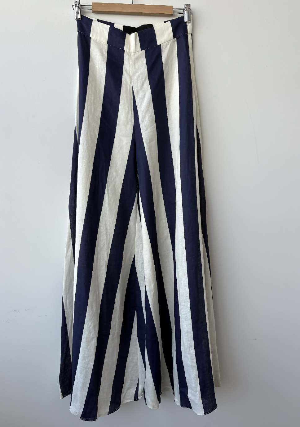 The EXCHANGE STORE Paper London Navy Stripe Wide Leg Pant, featuring bold stripes and a breezy linen finish, hangs on a wooden hanger against a crisp white background.