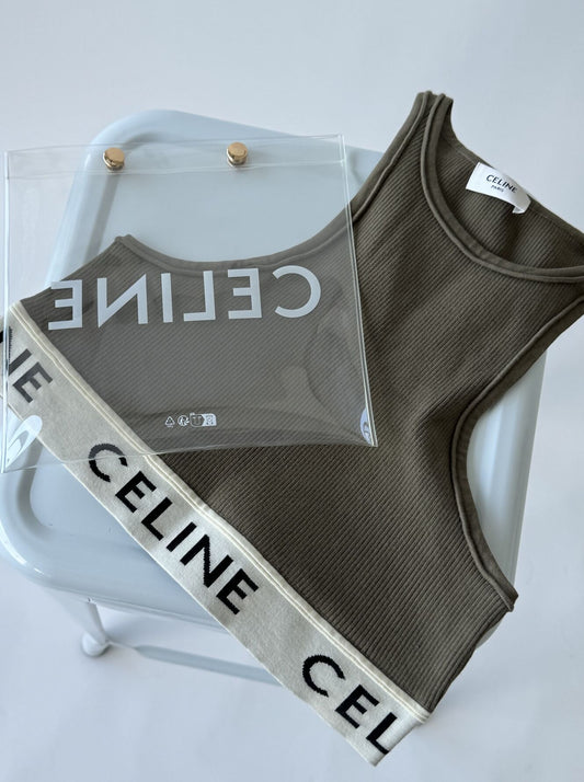 A Celine Jersey Top Bra by EXCHANGE, in ribbed olive green with a white logo band, is placed on a light gray chair. A transparent plastic bag labeled Celine rests atop the jersey top.