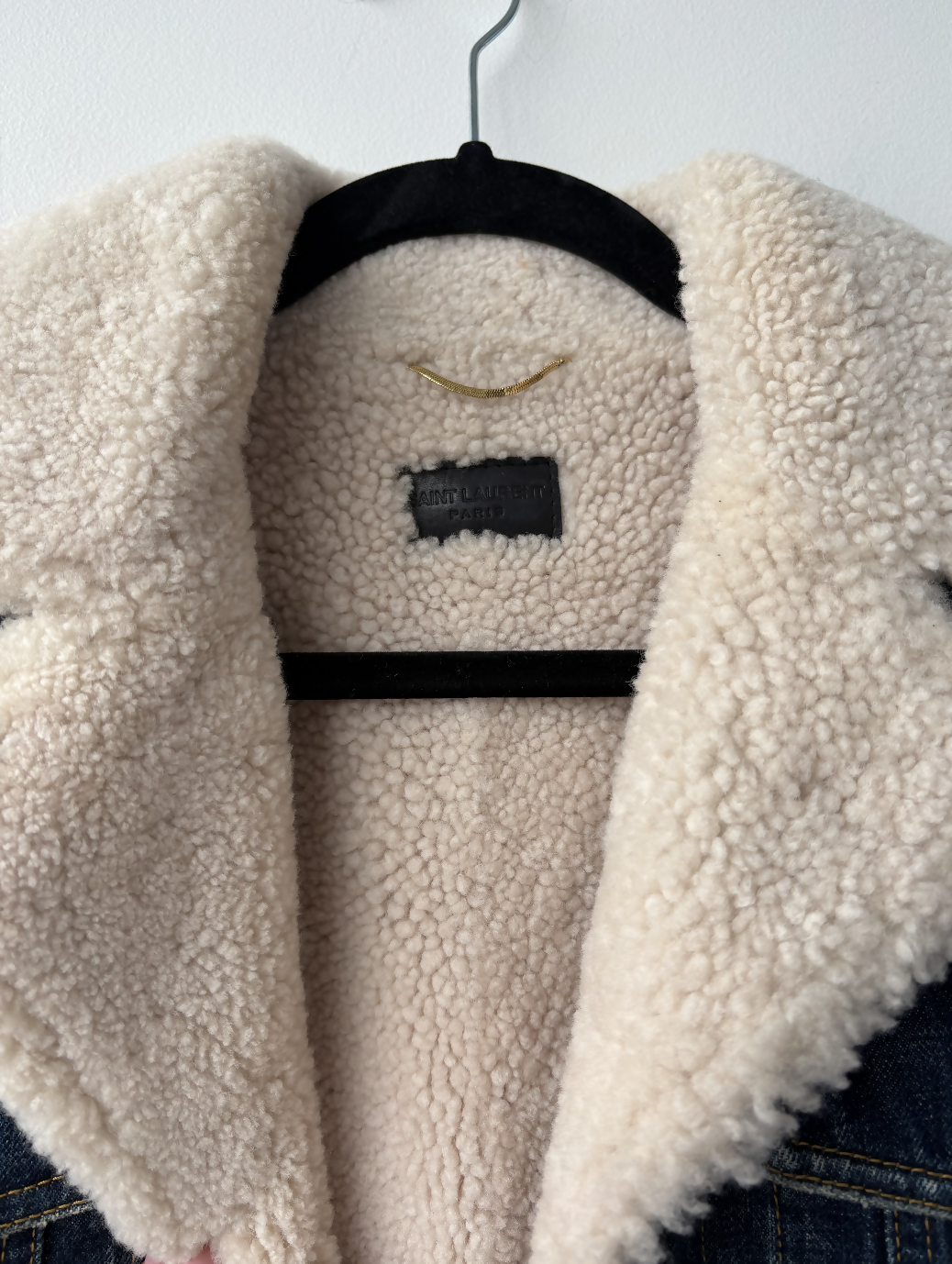 A close-up of the Yves Saint Laurent Paris Denim Shearing Jacket by EXCHANGE STORE, featuring a soft cream fleece lining and collar, displayed on a black velvet hanger against a white wall.