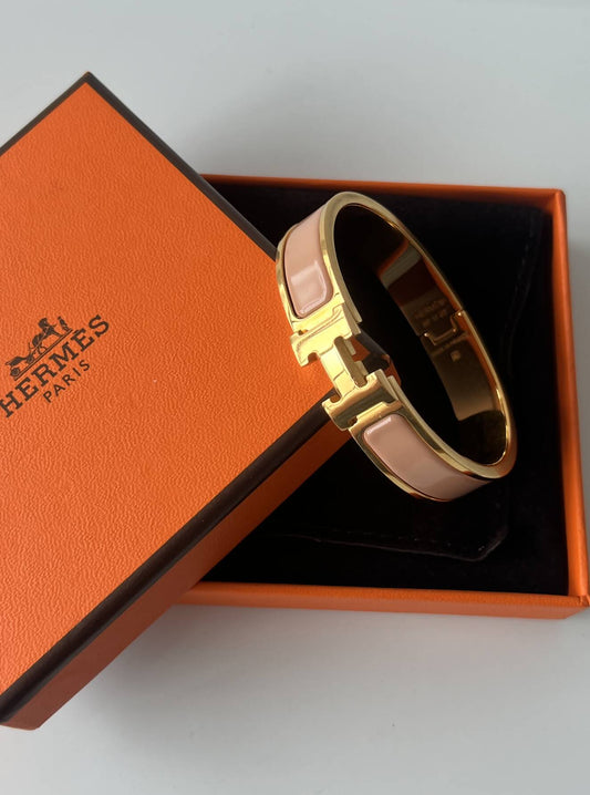 A flawless gold EXCHANGE Hermès Braclet - H braclet with light pink enamel details is shown resting on an orange Hermès box featuring black edges and the signature logo.