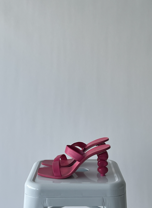 A brand new pair of EXCHANGE Cult Gaia Barbie Pink heels with unique rounded heels are displayed on a white plastic stool against a plain light gray background.