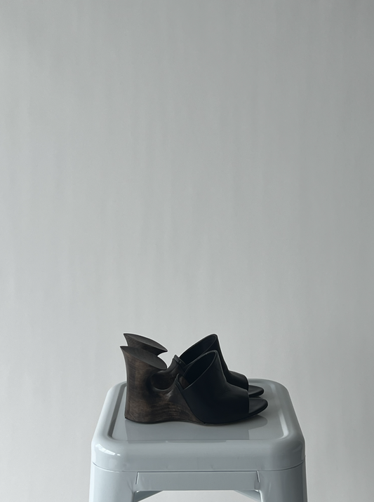 A pair of EXCHANGE Alaïa Cut Out Sculpture Mules - Noir sits on a light gray plastic stool against a plain, light gray background.