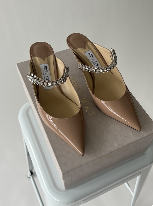 A pair of brand new Jimmy Choo Patent Mules by EXCHANGE with jeweled straps are displayed on a beige Jimmy Choo box atop a white stool, set against a plain background.
