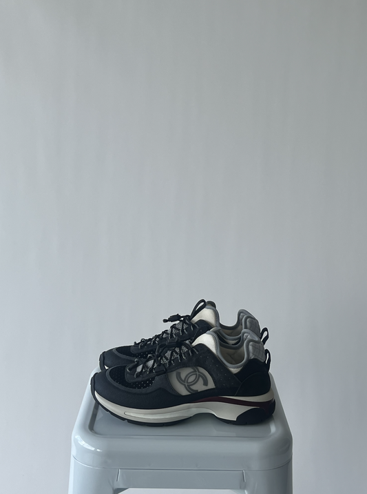 A pair of EXCHANGE Chanel Calf Skin Suede Sneakers in black and white, featuring a side logo, are neatly placed on a light gray stool against a plain white background.