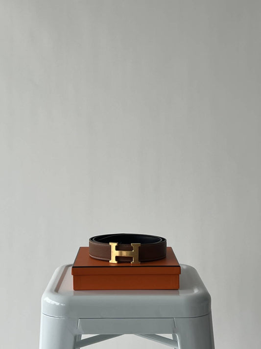 The EXCHANGE Hermès - Reversible Belt - Full Set features tan and black leather with a signature gold H buckle, elegantly rolled on an orange box atop a white stool against a light gray background.