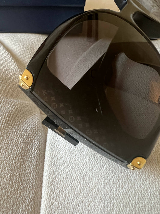 Close-up of Lulu Louis Vuitton Sunglasses with black gold hardware on the corners, subtle monogram lenses, resting on textured beige. Includes a case for safe storage.