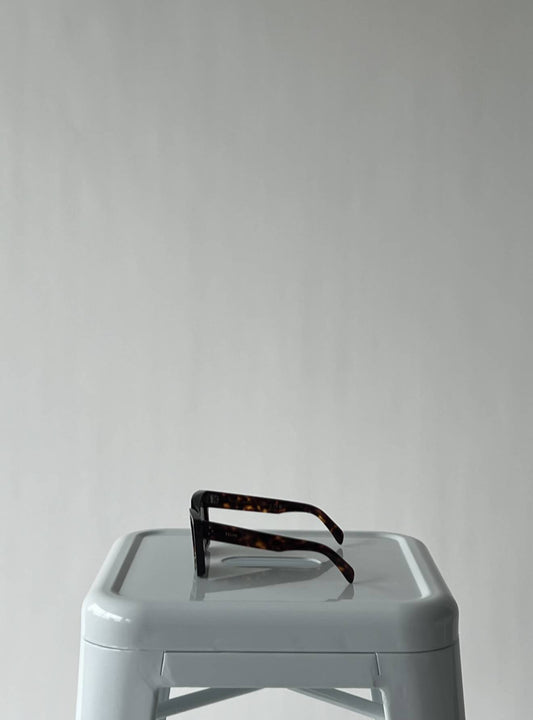 A pair of EXCHANGE Celine Glasses - CL40130I with an acetate frame rests upside down on a light gray plastic stool against a plain, neutral background.