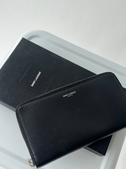 A black EXCHANGE Saint Laurent Wallet sits atop a matching box on a light gray surface. This stylish wallet offers multiple card slots for organized storage.